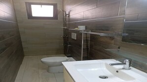 A bathroom with a toilet, a sink, a glass shower enclosure, and a small window.
