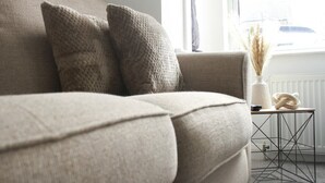 Un canapé beige agrémenté de deux coussins à motifs, une petite table d’appoint avec un vase blanc et une sculpture torsadée, et un radiateur en arrière-plan.