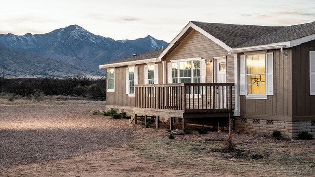 A single-story house with a wooden deck, large windows, and a mountain backdrop.