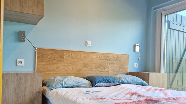 A bedroom with a wooden headboard, a bed with a patterned quilt, a nightstand, and a wall-mounted light. - Family home for 10 people, 600 m from the ocean – large garden (Mimizan)