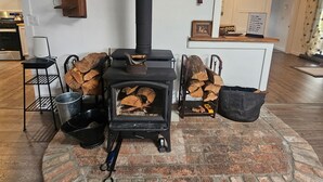 Un poêle à bois dans une pièce dont le sol est en briques, un support métallique contenant du bois de chauffage et un radiateur en bois fixé au mur.