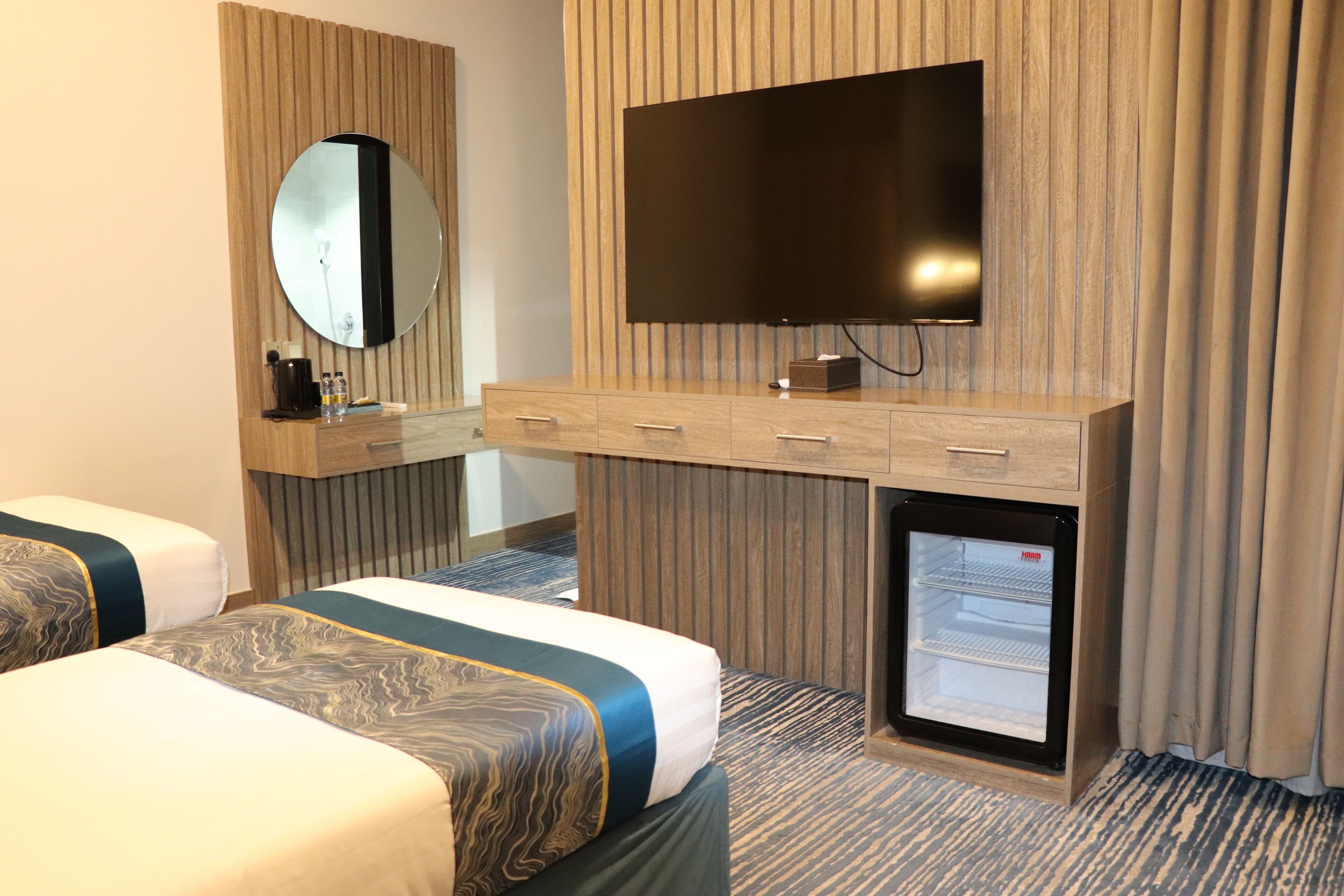 Ein Hotelzimmer mit zwei Betten, einem an einer Holzpaneele Wand montierten Fernseher, einer Minibar und einem kleinen Kühlschrank.