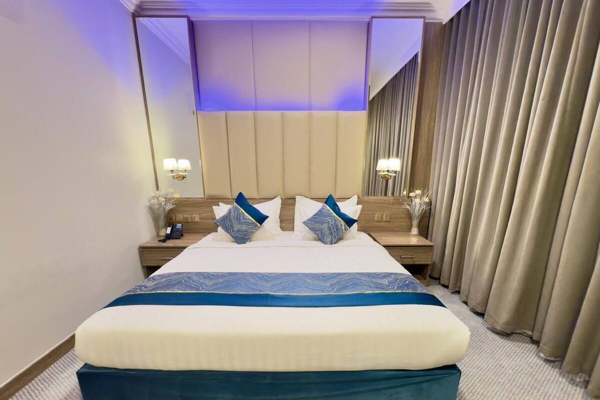 Une chambre d’hôtel avec un grand lit, deux tables de chevet avec des lampes, une tête de lit et un rideau sur le côté droit.