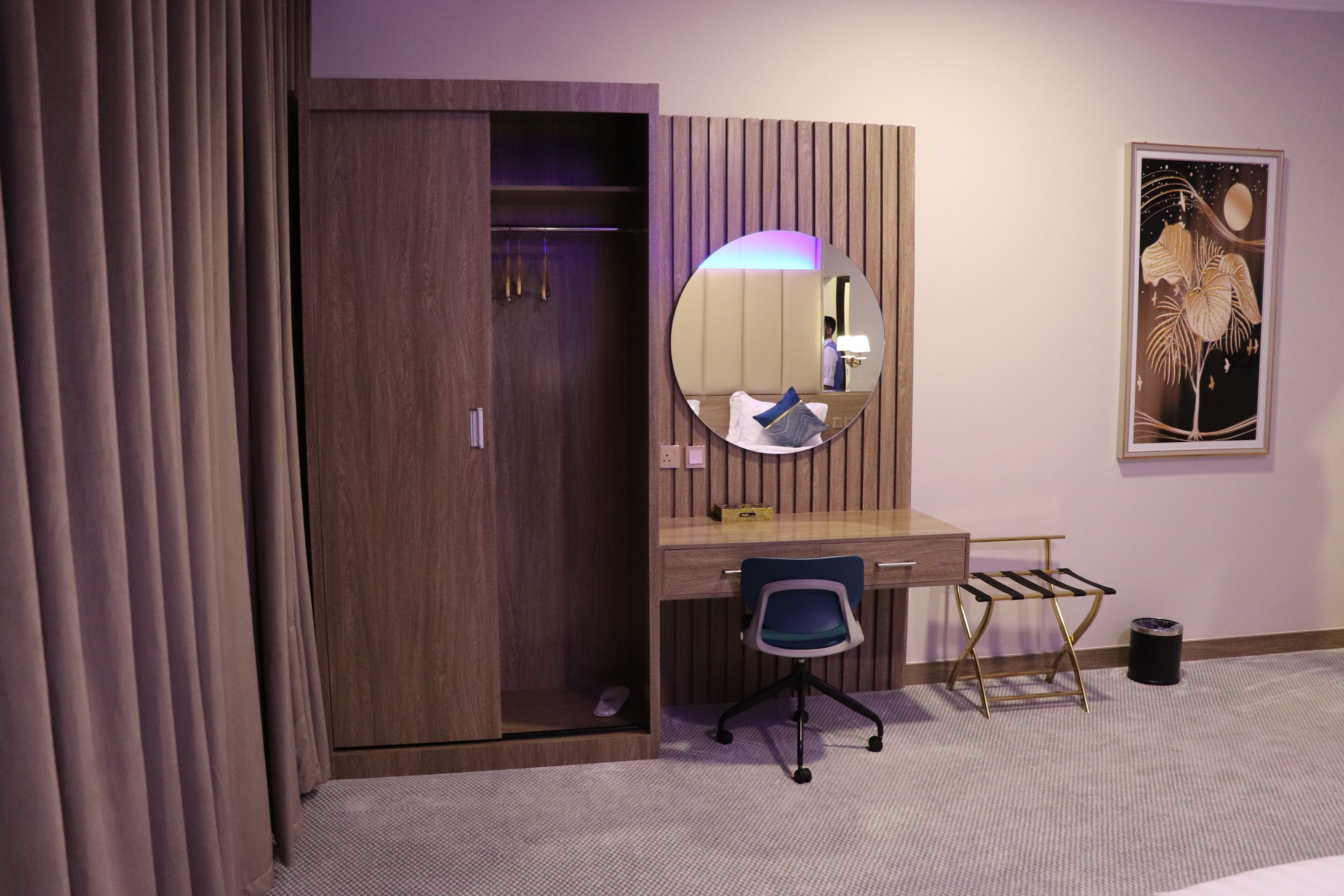Habitación de hotel con un armario de madera, un escritorio con silla, un espejo redondo y una obra de arte enmarcada en la pared.