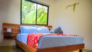 A wooden bed with a blue and red blanket, a nightstand with a lamp, and a window with a view of greenery.