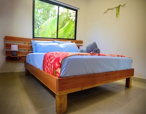 A wooden bed with a blue and red blanket, a nightstand with a lamp, and a window with a view of greenery.