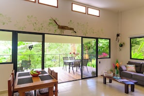 A modern living room with a dining area, large sliding glass doors to a balcony, and a sofa with a colorful cushion.