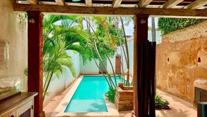 A pool area with a wooden deck, a bed, and a view of palm trees and a building in the background. - Paseo Colonial - Sabbatical 1 (Santo Domingo)