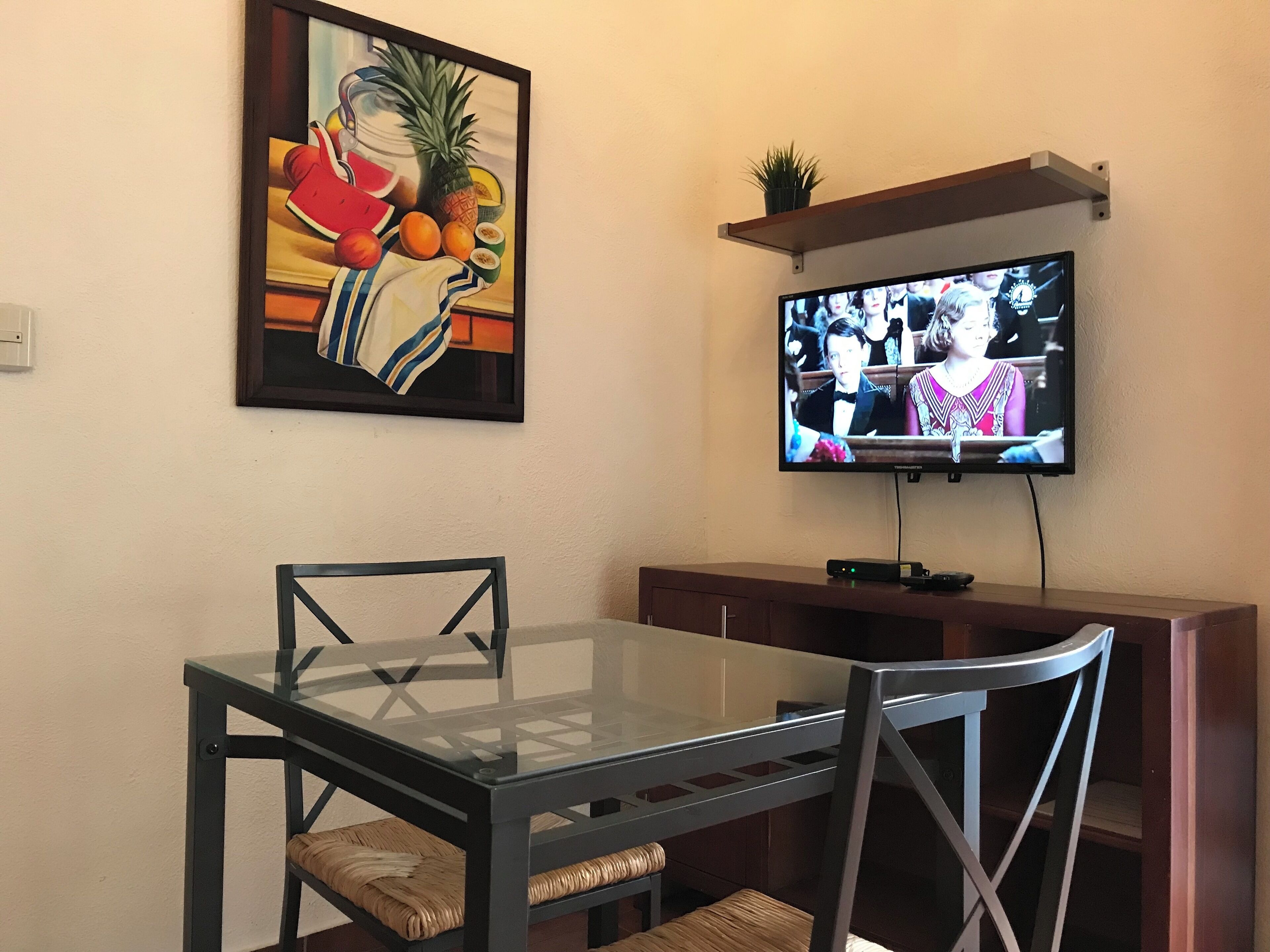 A dining area with a glass table and chairs, a painting of fruits, and a TV mounted on the wall.