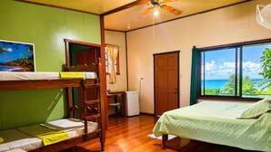 A room with bunk beds, a desk, a small refrigerator, a ceiling fan, and a large window with a view of the ocean.