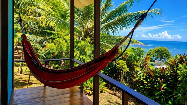 A hammock on a wooden deck with a view of the ocean and tropical vegetation.