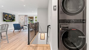 A modern laundry room with a large washer and dryer, a staircase with a railing, and a dining area with chairs and a table. - South Endzone Suite - 5 min walk to Lambeau, 4 BR/2 BA, Pool table, Foosball (Ashwaubenon)