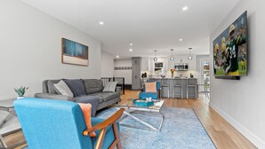 A modern living room with a sofa, a blue armchair, and a coffee table. There is a kitchen in the background with a dining area. - South Endzone Suite - 5 min walk to Lambeau, 4 BR/2 BA, Pool table, Foosball (Ashwaubenon)