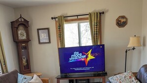 Een woonkamer met een televisie waarop het logo van 'The More You Know' staat, een bruine bank, een leren bank, een houten vloer en een grote klok.