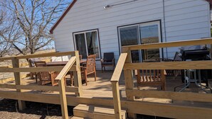Een houten terras met een tafel, stoelen, een barbecue en een wit gebouw met grote ramen.
