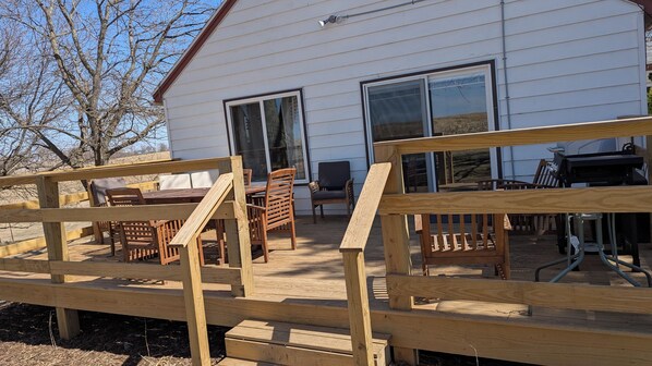 A wooden deck with a table and chairs, a grill, and a white building with large windows. - Private, Cozy, Driftless 2BR Farmhouse on 80 acres, 2 miles from Viroqua, WI (Viroqua)