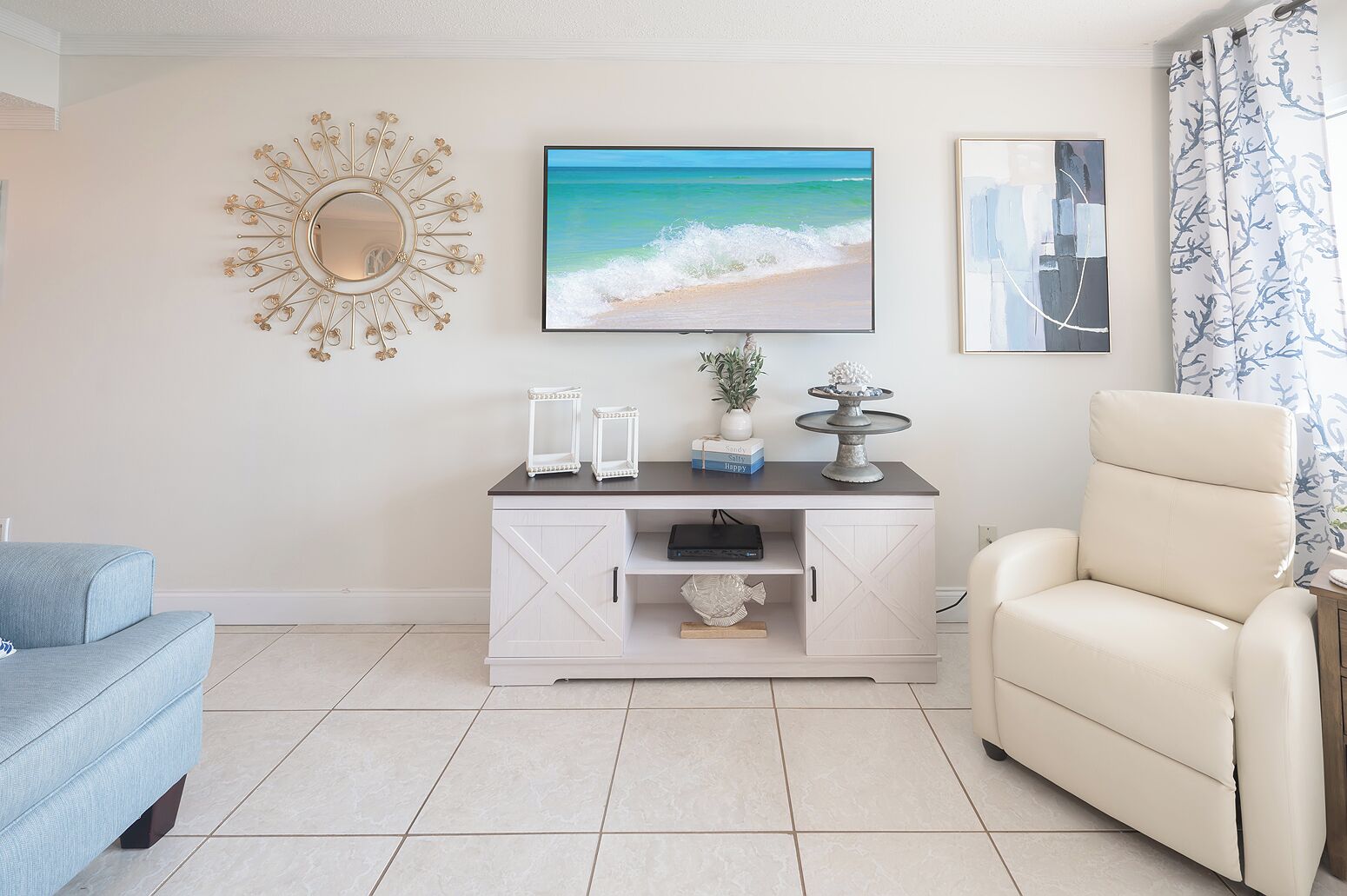 A living room with a white cabinet, a blue sofa, a beige armchair, a TV displaying a beach scene, and a decorative mirror.