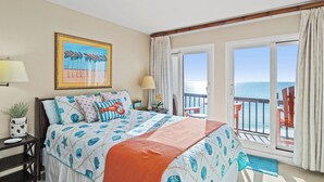 Habitación de hotel con cama, un cuadro, una mesita de noche, lámpara, ventana con vista al mar y balcón con sillas.