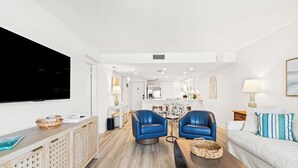 A modern living room with a flat-screen TV, blue leather chairs, a white sofa, and a dining area with a white table and chairs. - Pinnacle Port, C1-401 (Panama City Beach)
