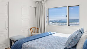 A bedroom with a large window offering a view of the ocean, a bed with blue bedding, a chair, and white walls.