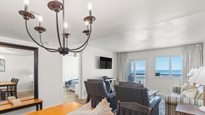 A hotel room with a wooden dining table, a chandelier, a sofa, and a view of the ocean through large windows.