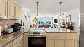 A spacious kitchen with granite countertops, a built-in oven, and a large island. There are pendant lights, a dining area with a view, and a living room with a fireplace. - Pinnacle Port, C3-604 (Panama City Beach)