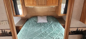 A compact RV interior with a bed, wooden cabinets, a window, and a thermostat.