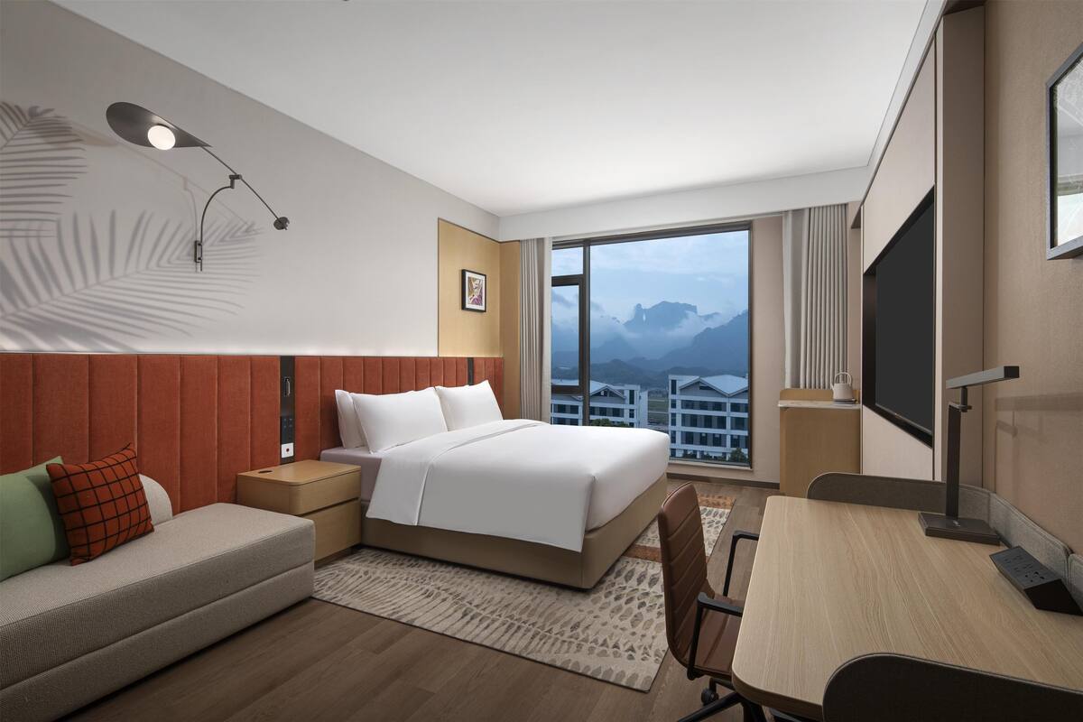 Habitación de hotel con cama, sofá, escritorio y un amplio ventanal con vistas a la montaña.