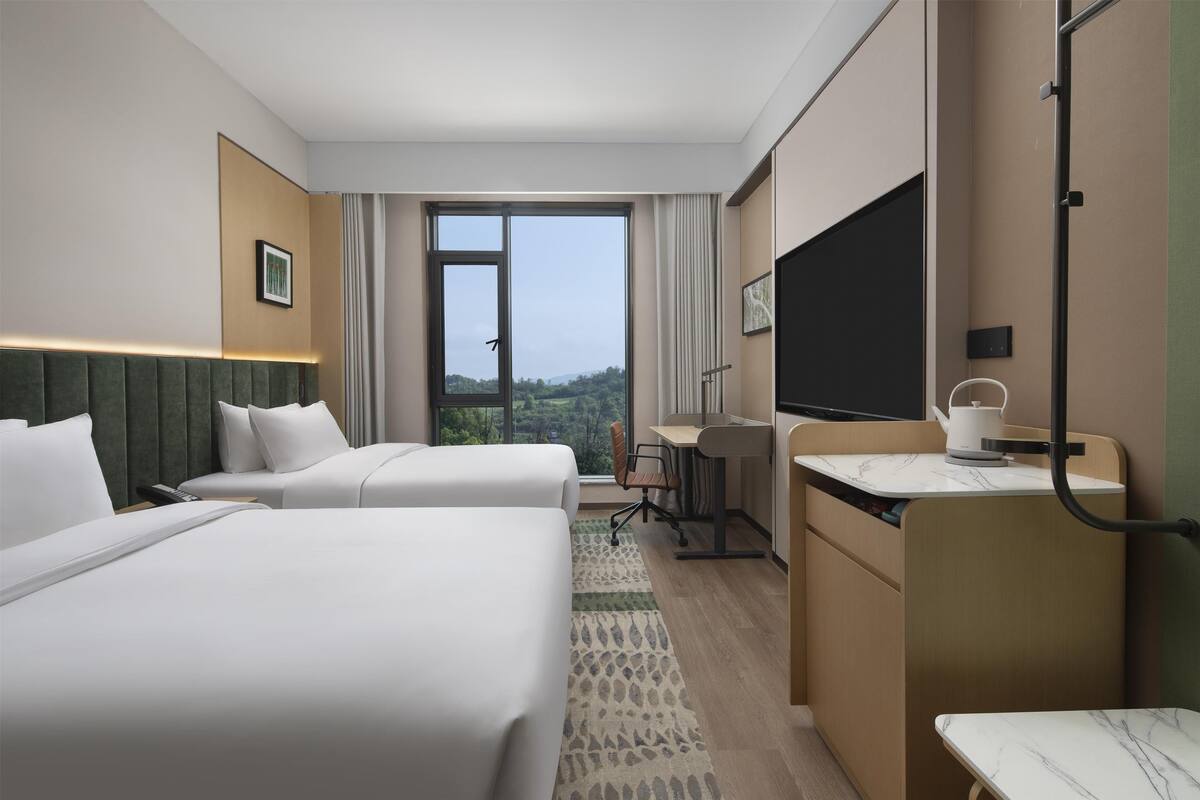 Habitación de hotel con dos camas, un escritorio, una silla, un televisor y un amplio ventanal con vistas al exterior.