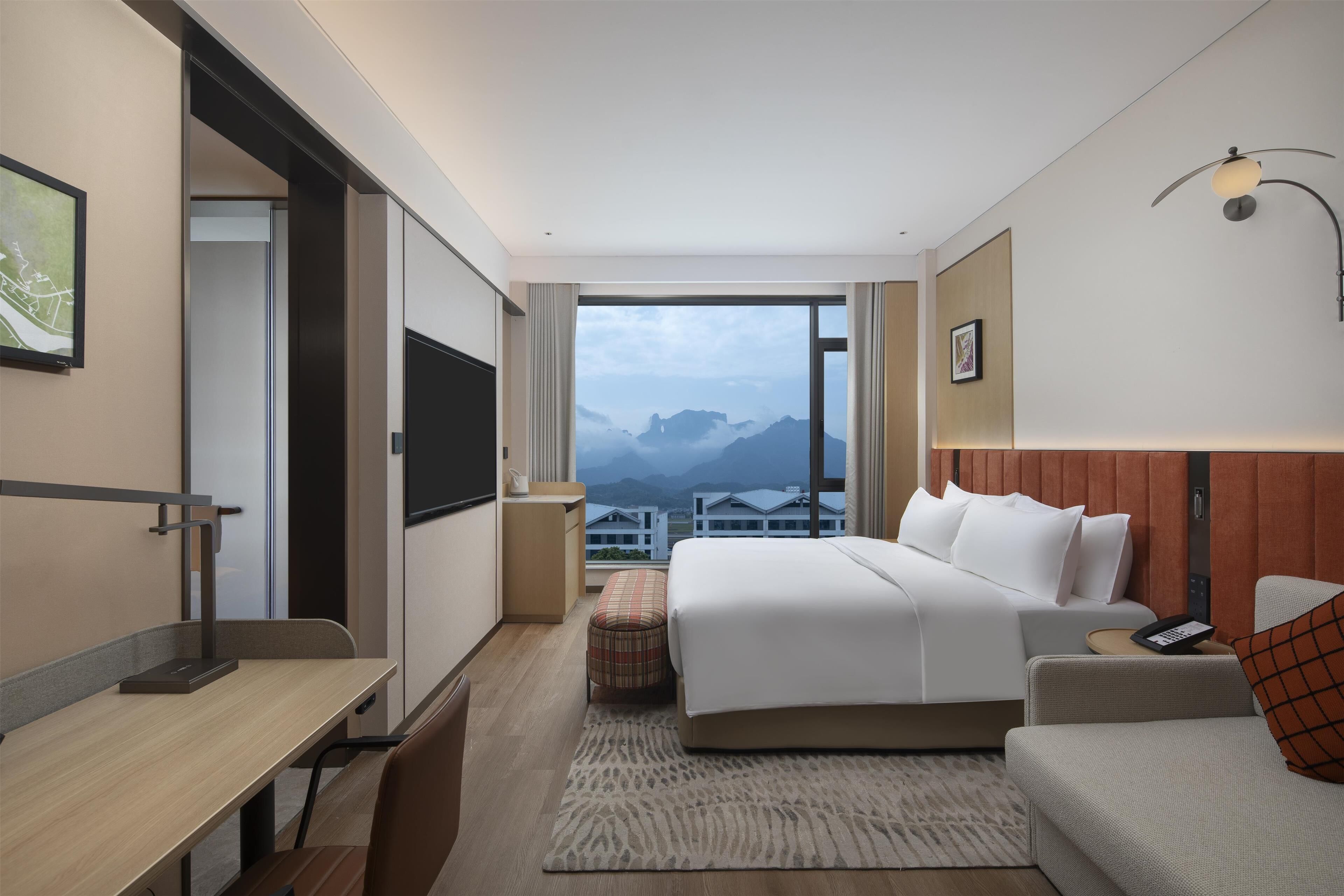 A modern hotel room with a large bed, a desk, a chair, a sofa, and a view of mountains.