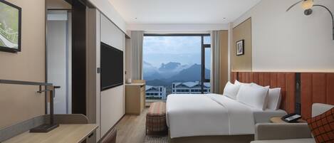 A modern hotel room with a large bed, a desk, a chair, a sofa, and a view of mountains.
