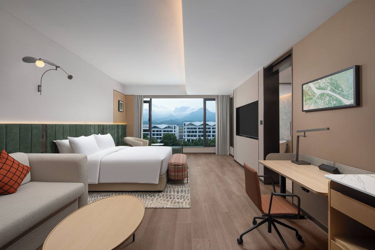 Una habitación de hotel moderna con cama, escritorio, sofá y vistas a montañas a través de amplios ventanales.