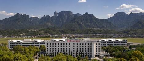 Ein modernes Hotel mit flachem Dach und mehreren Etagen, umgeben von üppigem Grün und Bergen im Hintergrund.