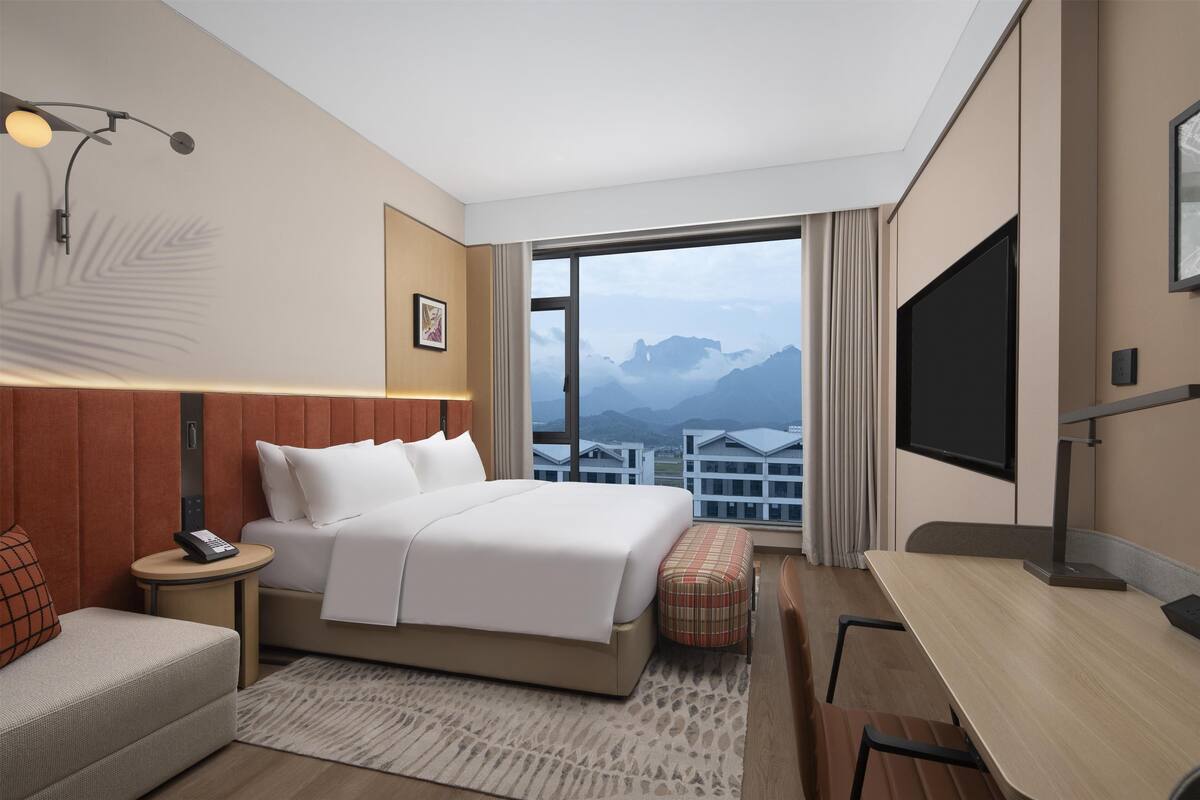 Habitación de hotel con cama, escritorio, silla, pequeña mesa, teléfono, televisor y ventana grande con vistas a la montaña.