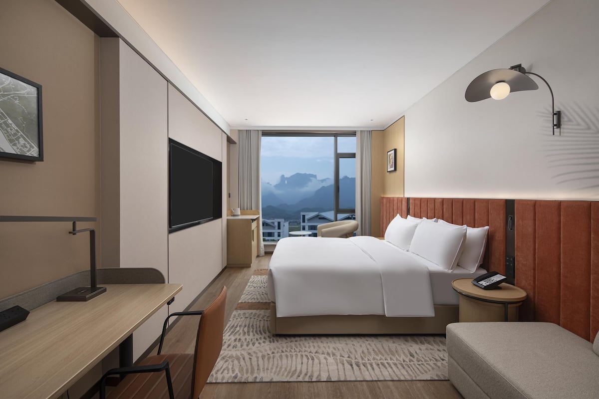 Habitación de hotel con cama, escritorio, silla, mesita de noche, lámpara y vista a las montañas por la ventana.
