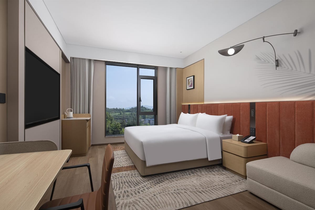 Habitación de hotel con cama, escritorio, silla, mesita de noche, lámpara y vistas al exterior.