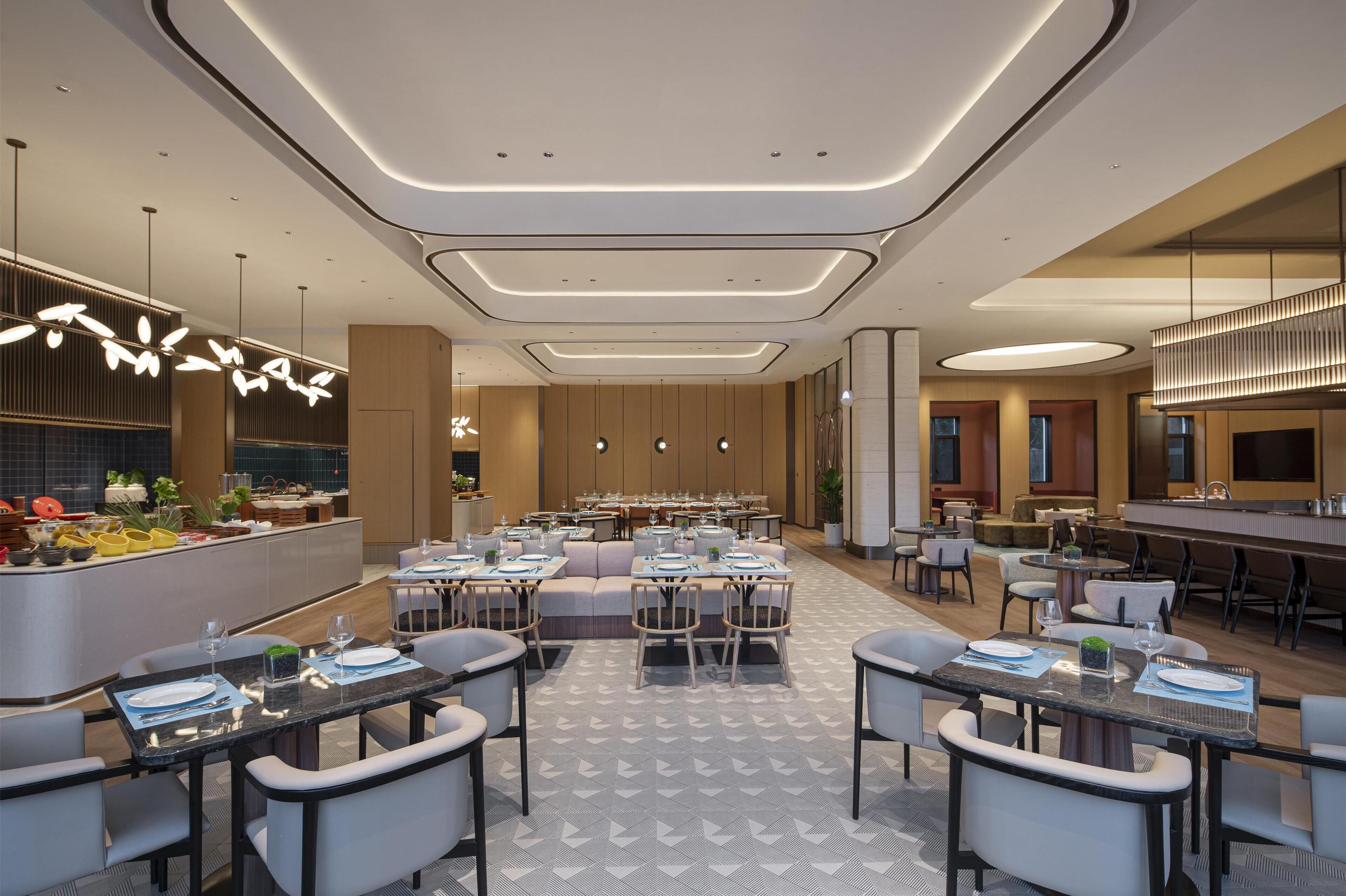 Un espace de restauration moderne dans un hôtel, offrant de nombreuses options de sièges, notamment des tables et des banquettes, ainsi qu’un bar central.