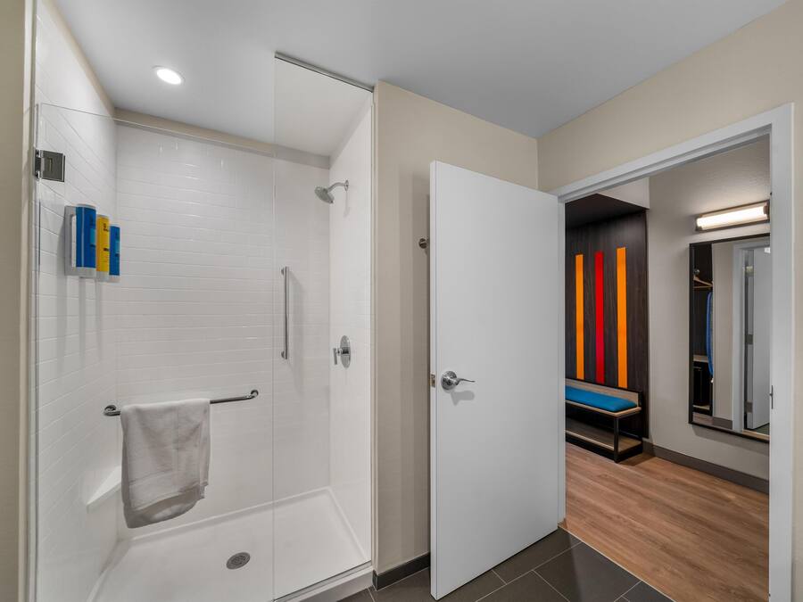 Un baño moderno con cabina de ducha de vidrio, una bañera blanca y una puerta que da a un pasillo con un banco y un espejo.