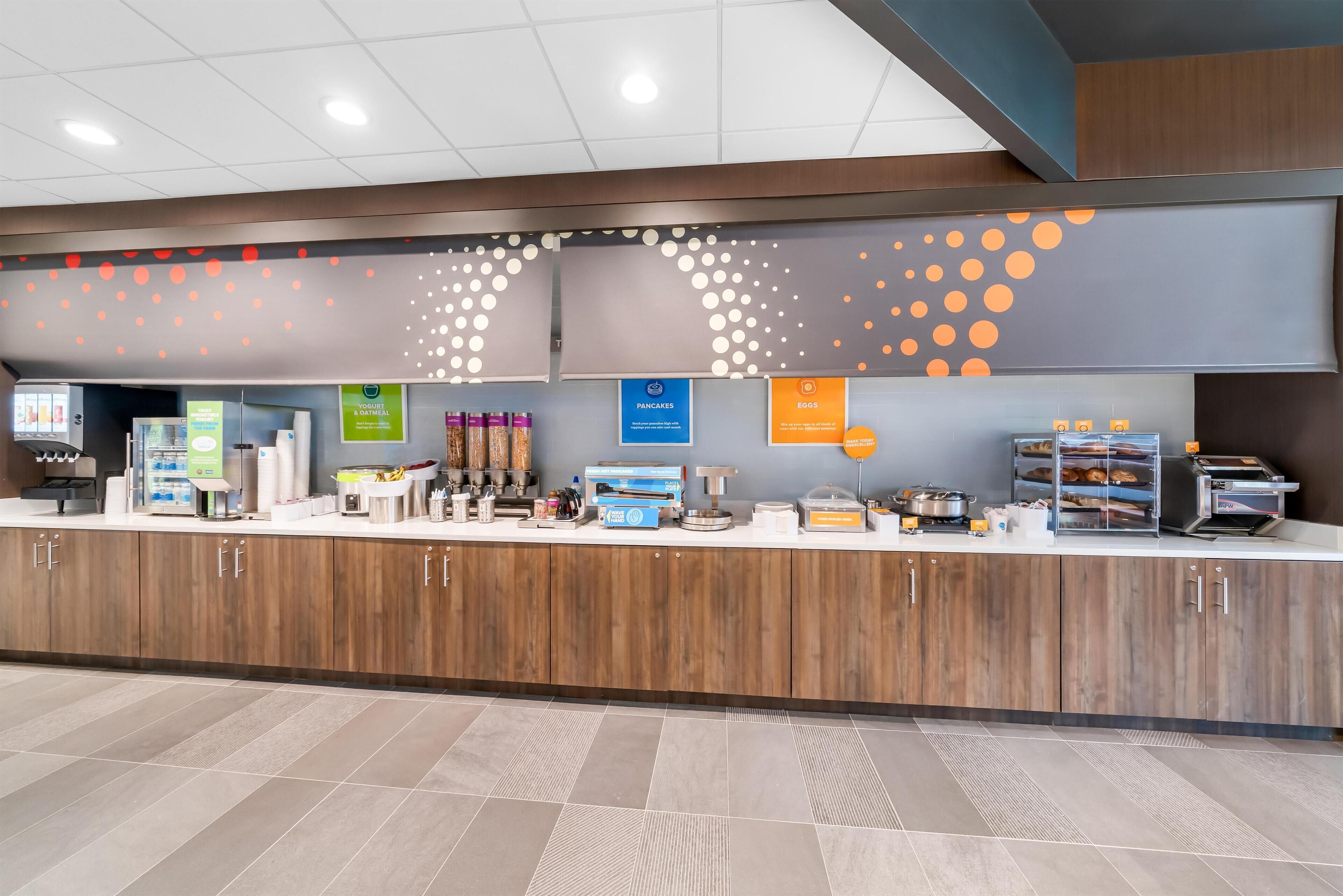 A modern breakfast area with a wooden counter, various food items, and signs indicating available breakfast options.