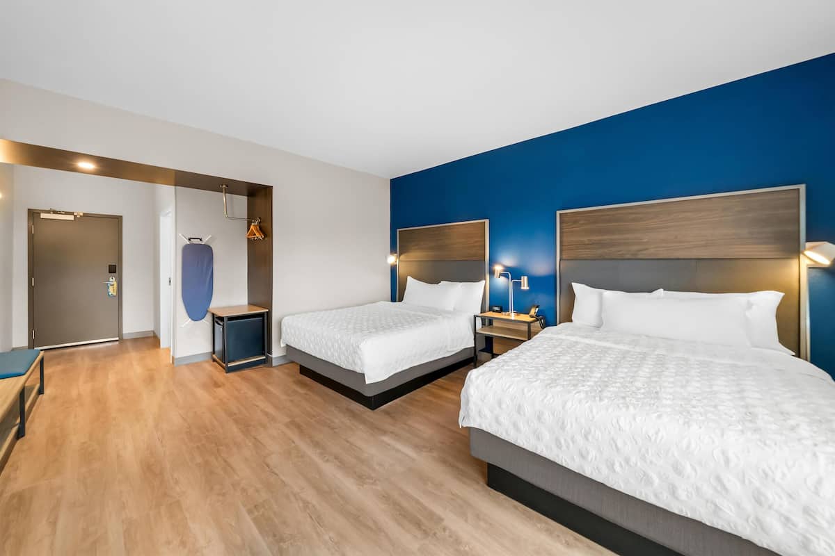 Quarto de hotel com duas camas, piso de madeira, parede azul e um pequeno espaço para sentar.