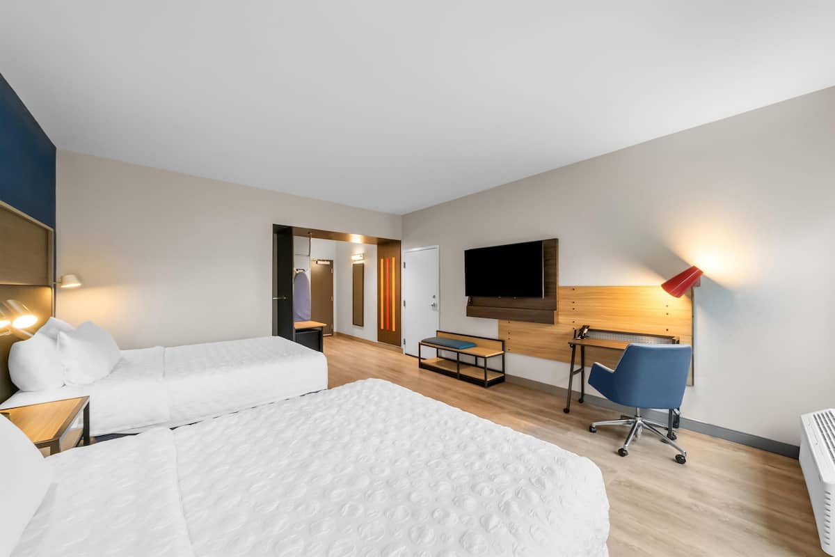 Quarto de hotel com duas camas, uma escrivaninha, uma cadeira, uma TV e um abajur vermelho.
