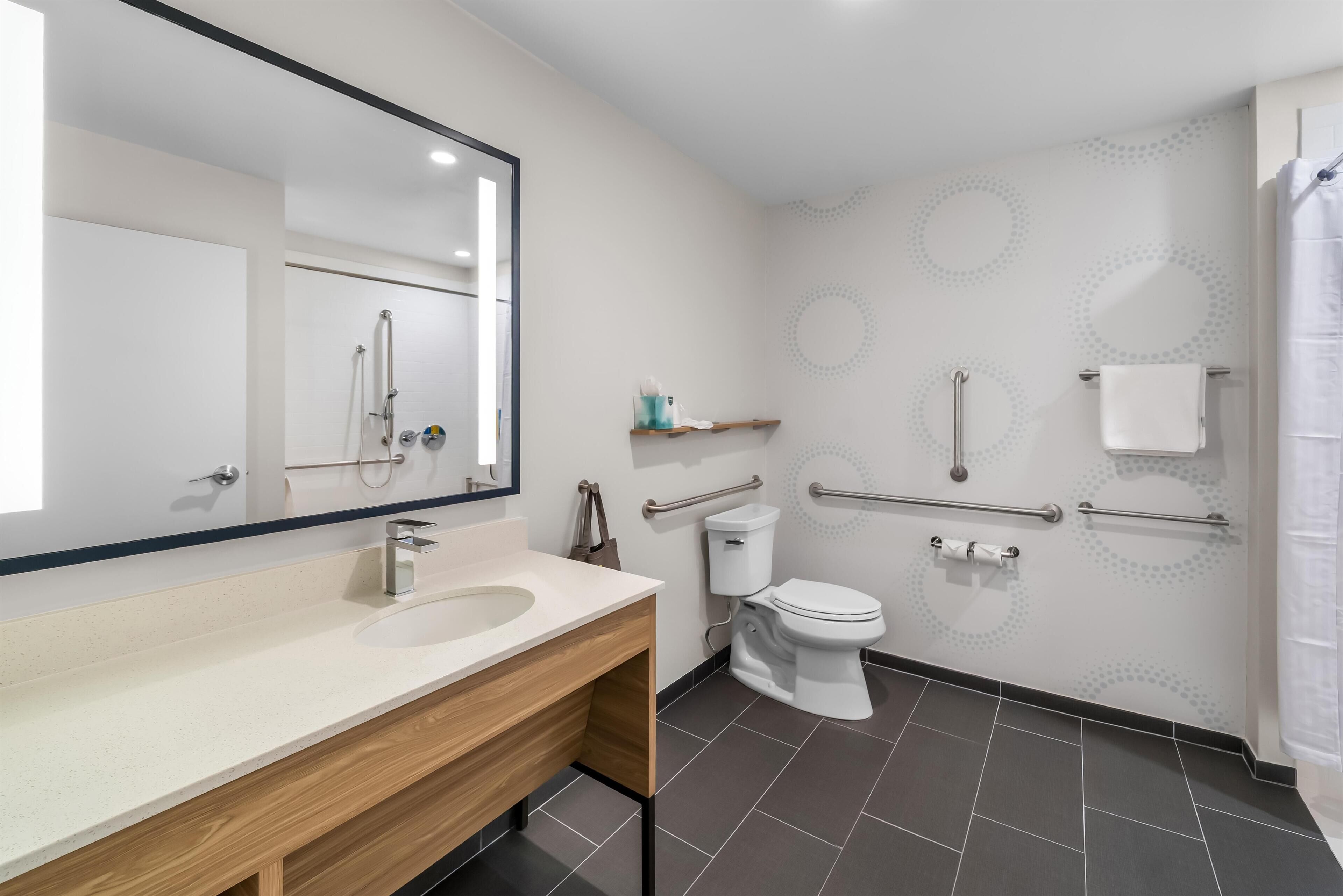 Un baño moderno con un espejo grande, un mueble de tocador de madera, un lavabo blanco, un inodoro y una zona de ducha.