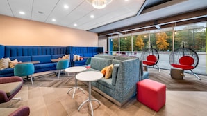 Een moderne lounge met een blauwe bank, blauwgroene fauteuils en rode stoelen. Er staan witte tafels en een rode poef. De ruimte heeft grote ramen met uitzicht op bomen en een parkeerplaats.