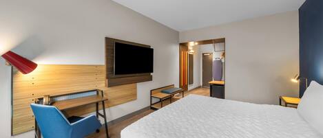 A hotel room with a bed, a desk with a chair, a TV mounted on the wall, and a mirror.