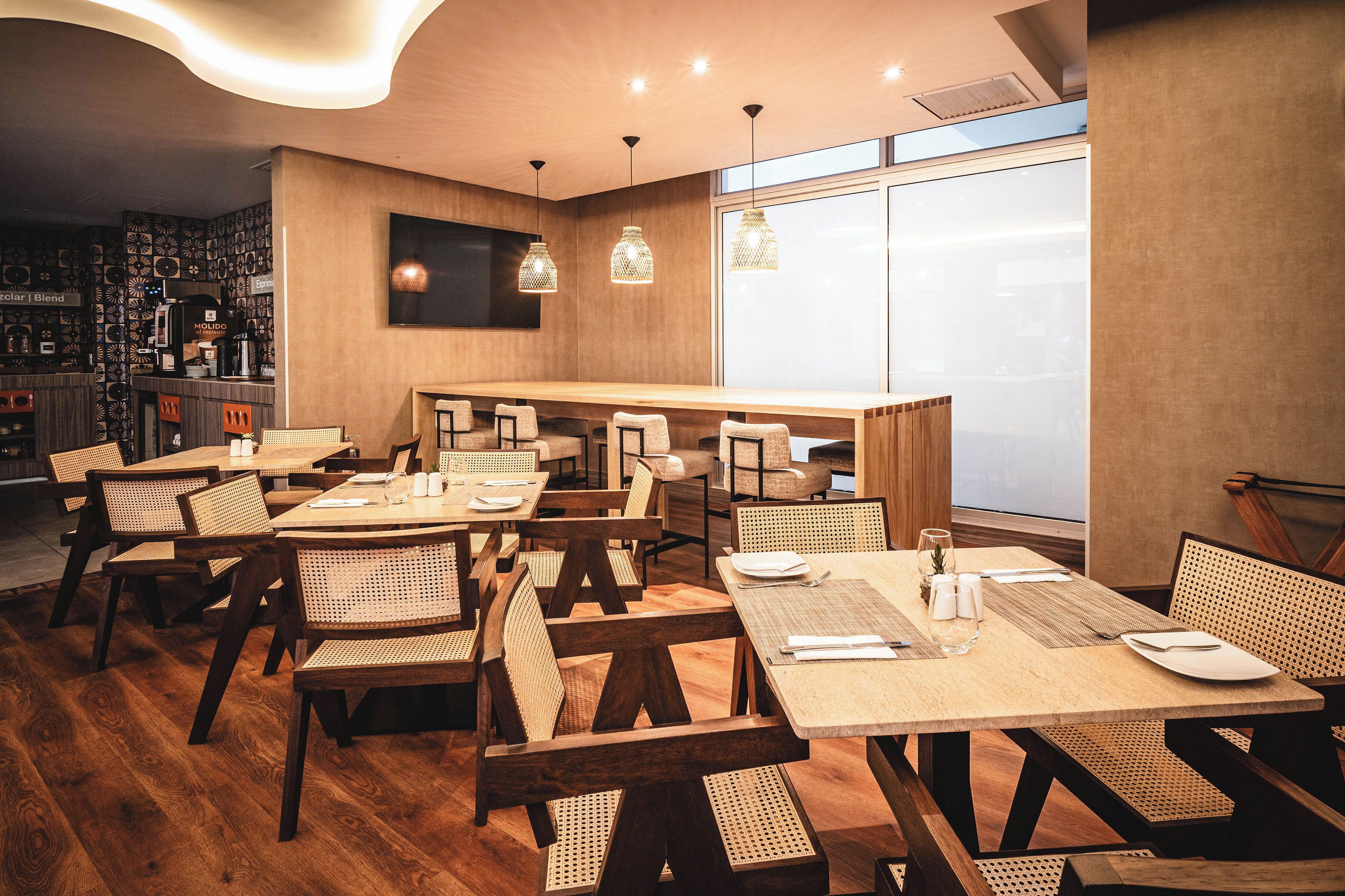 Un restaurante con mesas y sillas de madera, una zona de bar y un amplio ventanal que proporciona luz natural.