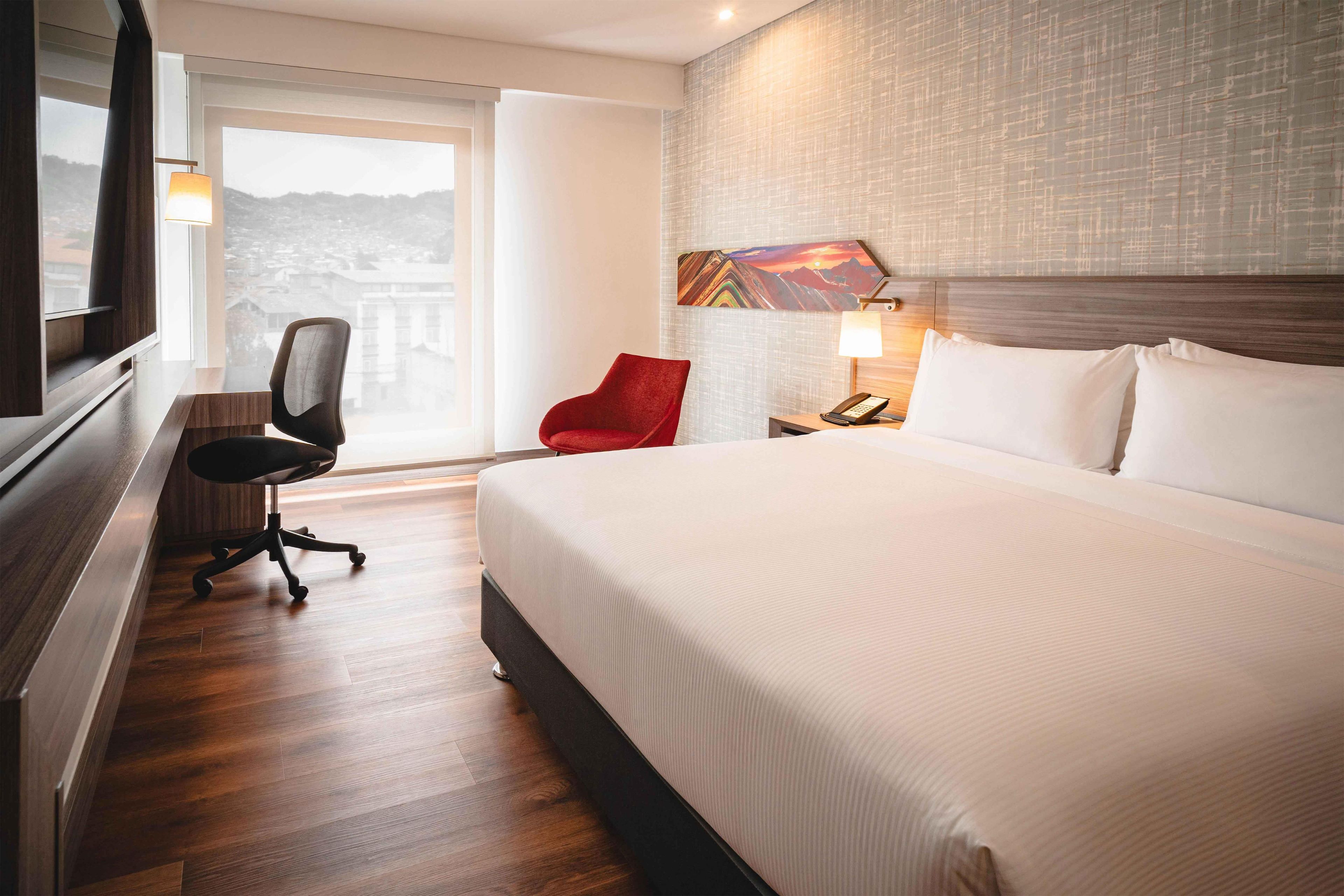 A hotel room with a large bed, a desk with a chair, a nightstand with a lamp, and a painting on the wall.