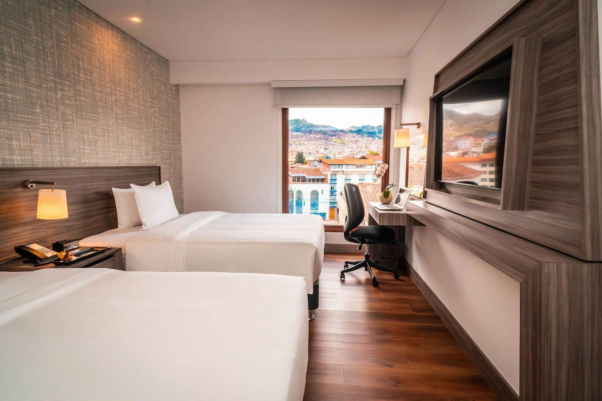 A hotel room with two beds, a desk, a chair, and a large window with a city view.