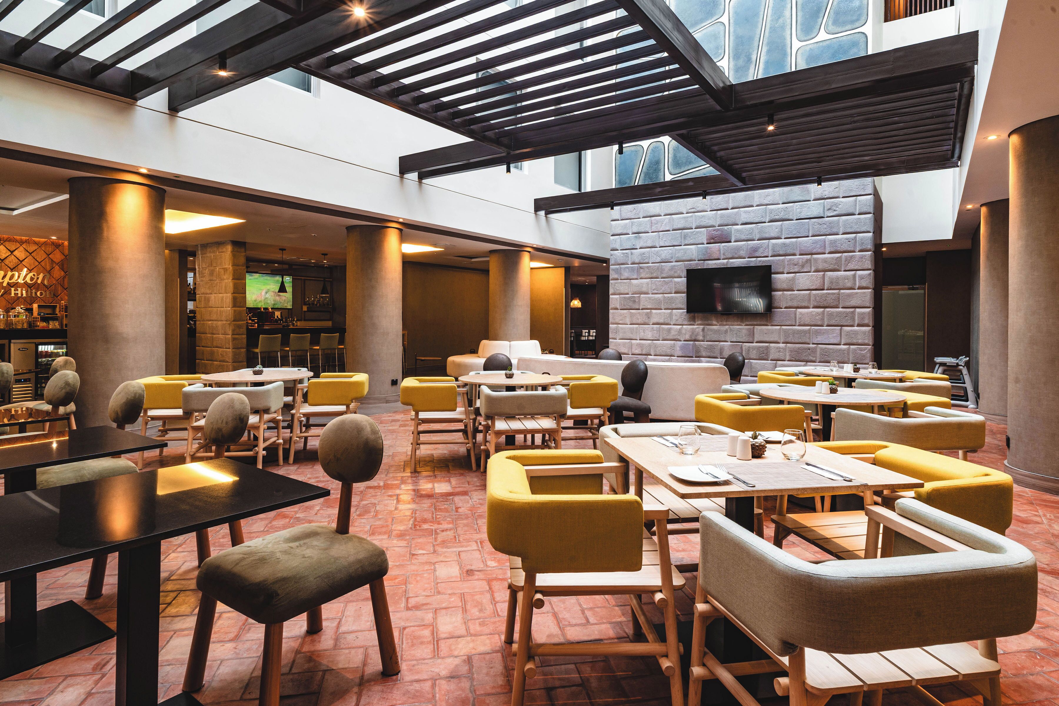 A spacious indoor dining area with high ceilings, exposed beams, and a brick accent wall. Tables and chairs are arranged for dining.