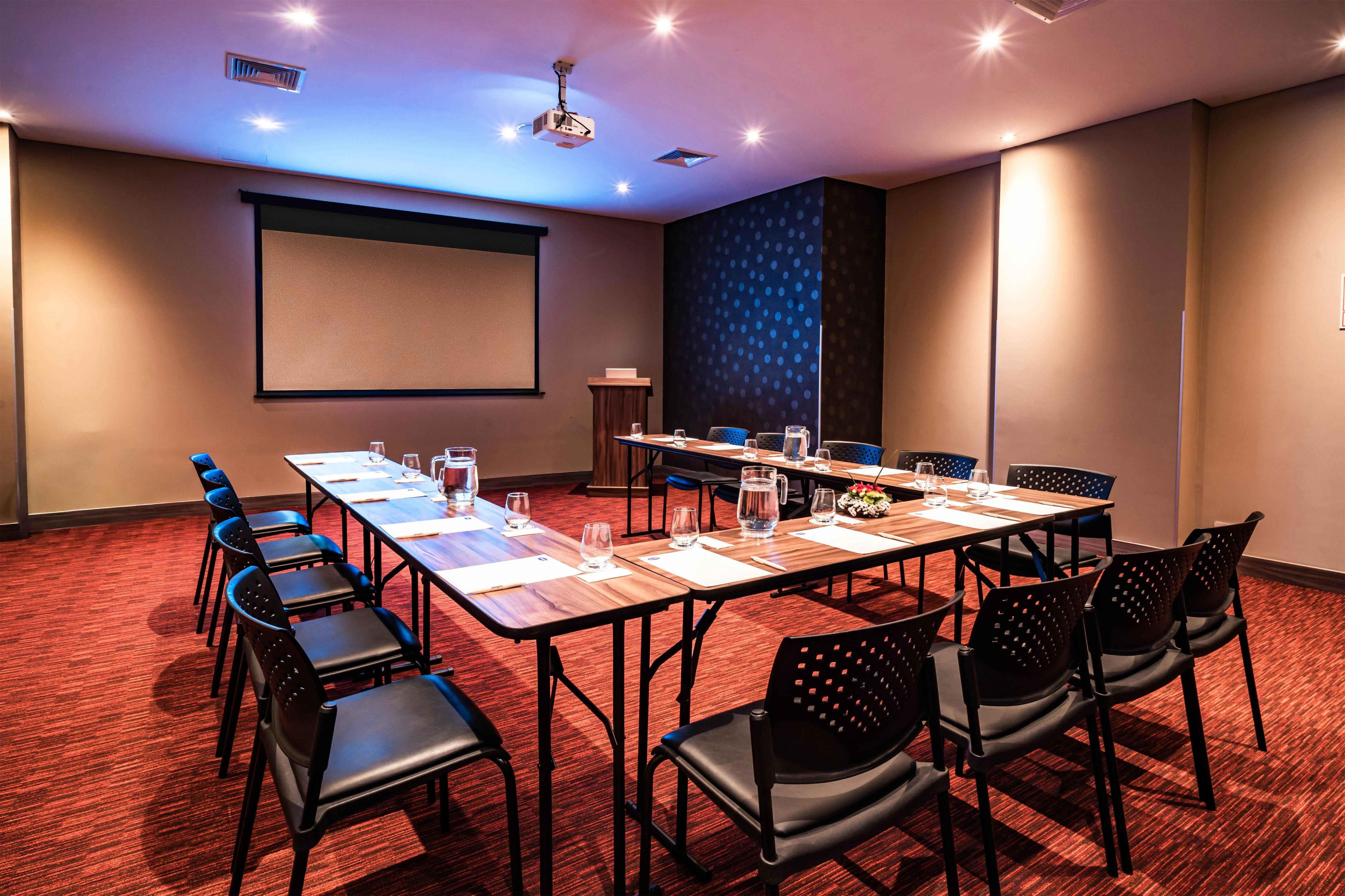 A conference room with a projector screen, a large table with chairs, and a carpeted floor.