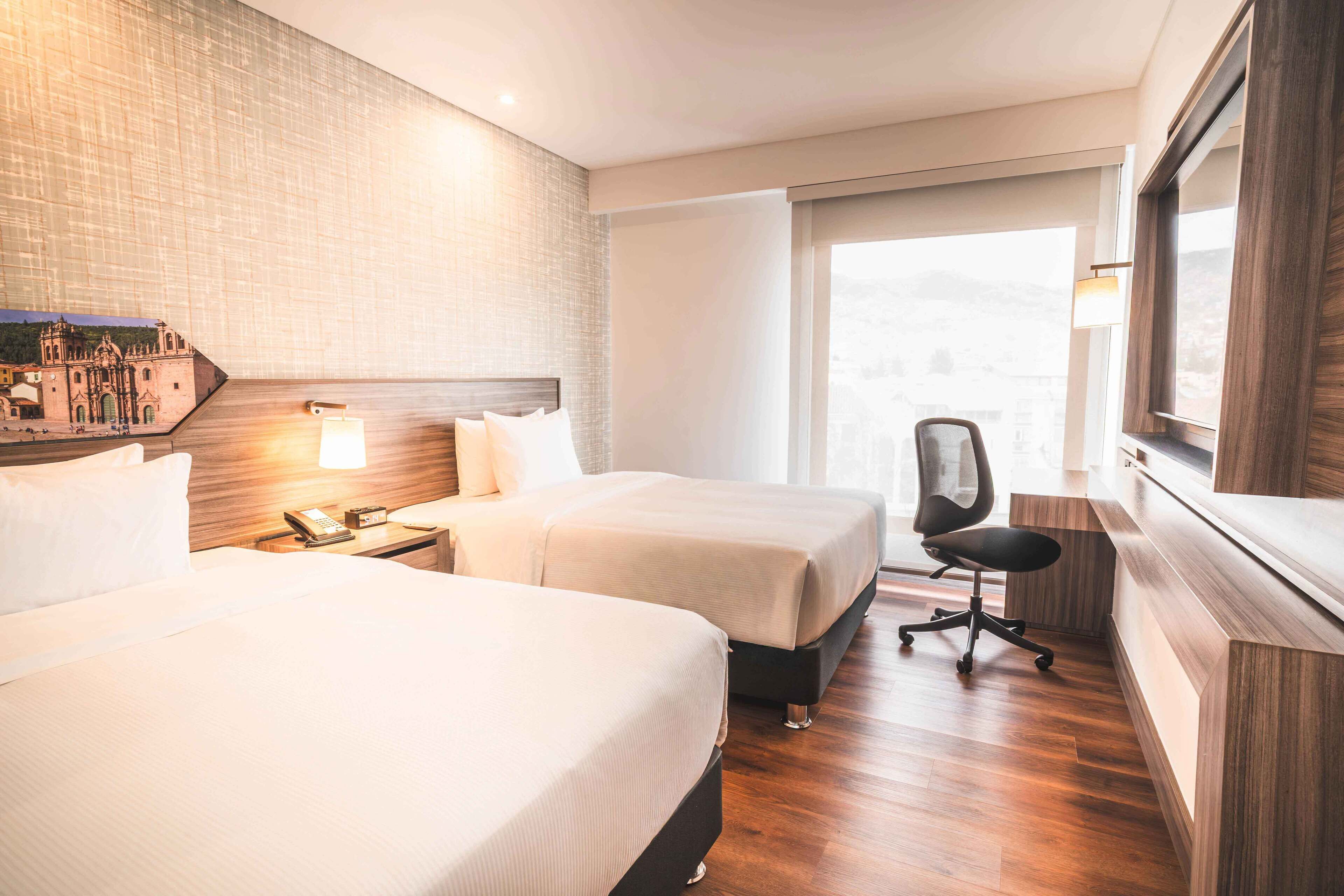 A hotel room with two beds, a desk, a chair, and a large window with a view.